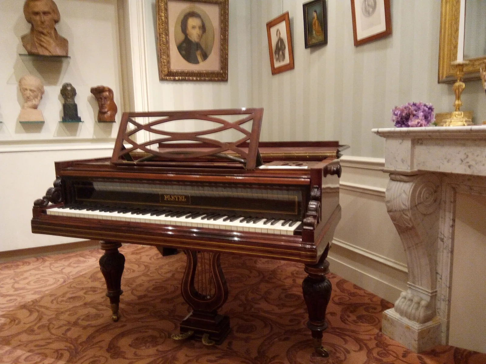 Lesklé křídlo Pleyel s vyřezávaným stojanem pedálů ve tvaru lyry v obloženém salonku, na zdi za ním zarámované portréty